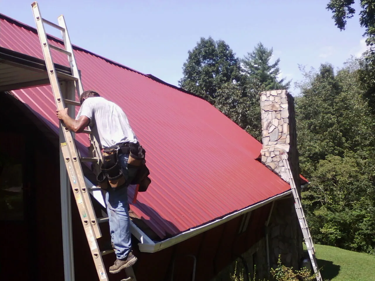 Roofer on a ladder beside a red metal roof and stone chimney — Soffit & Fascia Repair by Roofing Brothers in Champlin